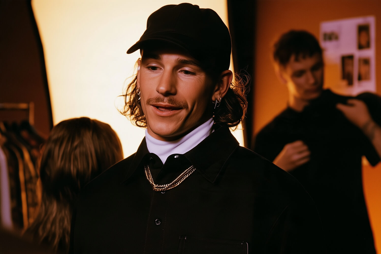 Man in a black cap and white turtleneck smiling backstage as a crew member works behind him